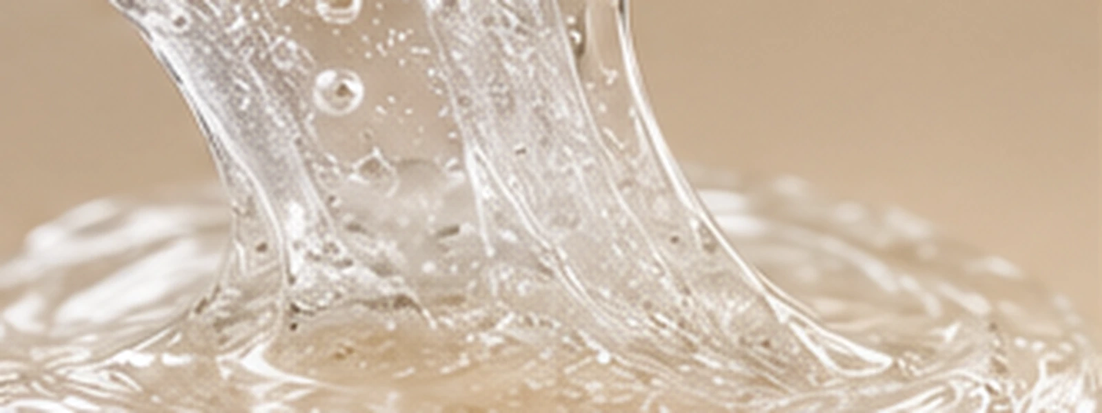 Clear elastic viscous skincare texture stretching across a warm ivory surface.