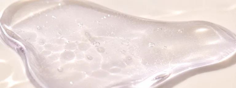 Cool pearl-lilac translucent serum with tiny suspended microbubbles on warm ivory.